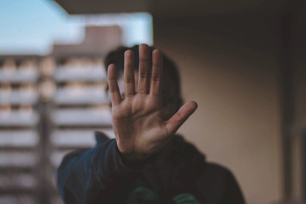 A person holds up their hand towards the camera, palm open and fingers spread, obscuring their face. The background appears to be a blurred urban setting with out-of-focus buildings.