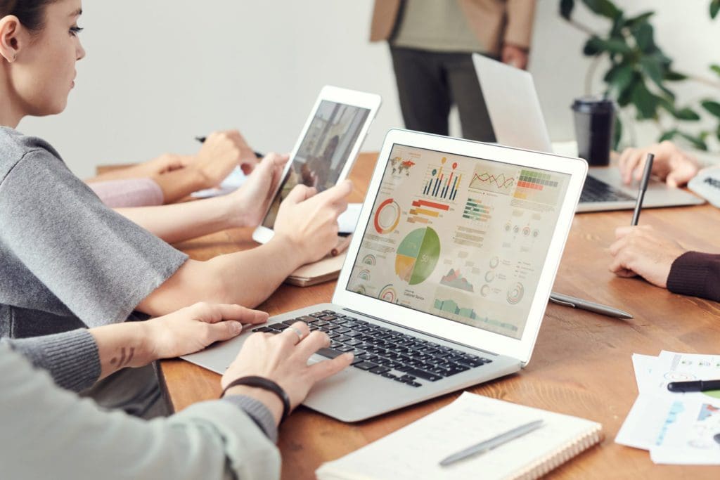 A group of people is seated around a wooden table working on electronic devices. Some are using laptops while others are using tablets. The screens show colorful charts and graphs. Notebooks and papers are also on the table, suggesting a meeting or collaborative work session.