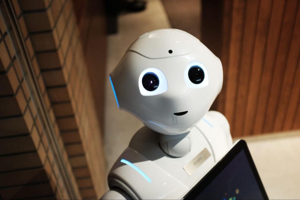 A white humanoid robot with large, round black eyes and a small mouth is looking up. The robot has a tablet attached to its body and is illuminated by soft lighting. The background features a brick wall and wooden paneling.