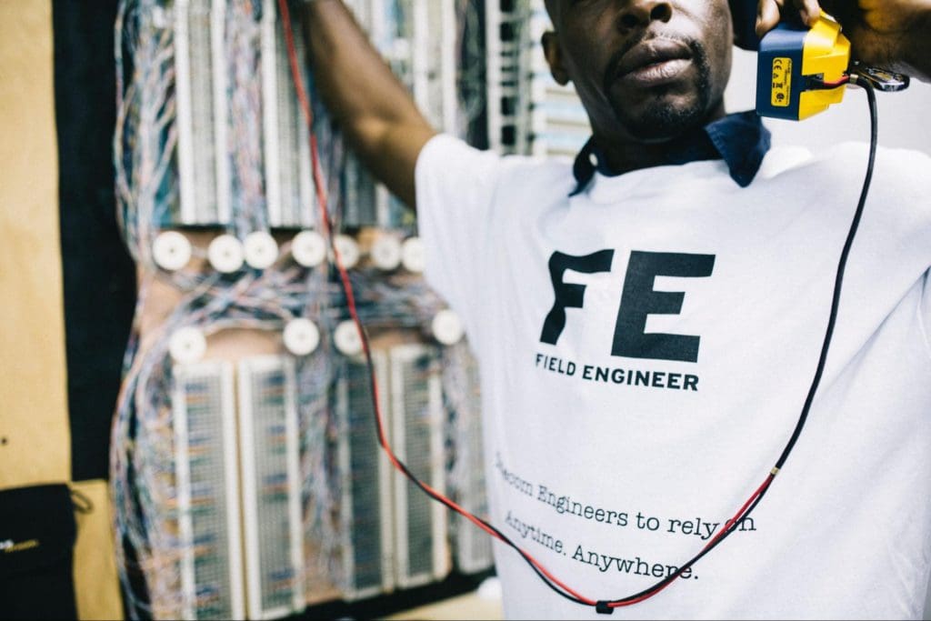 A person wearing a "Field Engineer" t-shirt is holding a yellow device while working on a complex set of multiple colored wires in an electrical panel. The text on the shirt reads, "Field Engineer. For Engineers to rely on Anytime. Anywhere.
