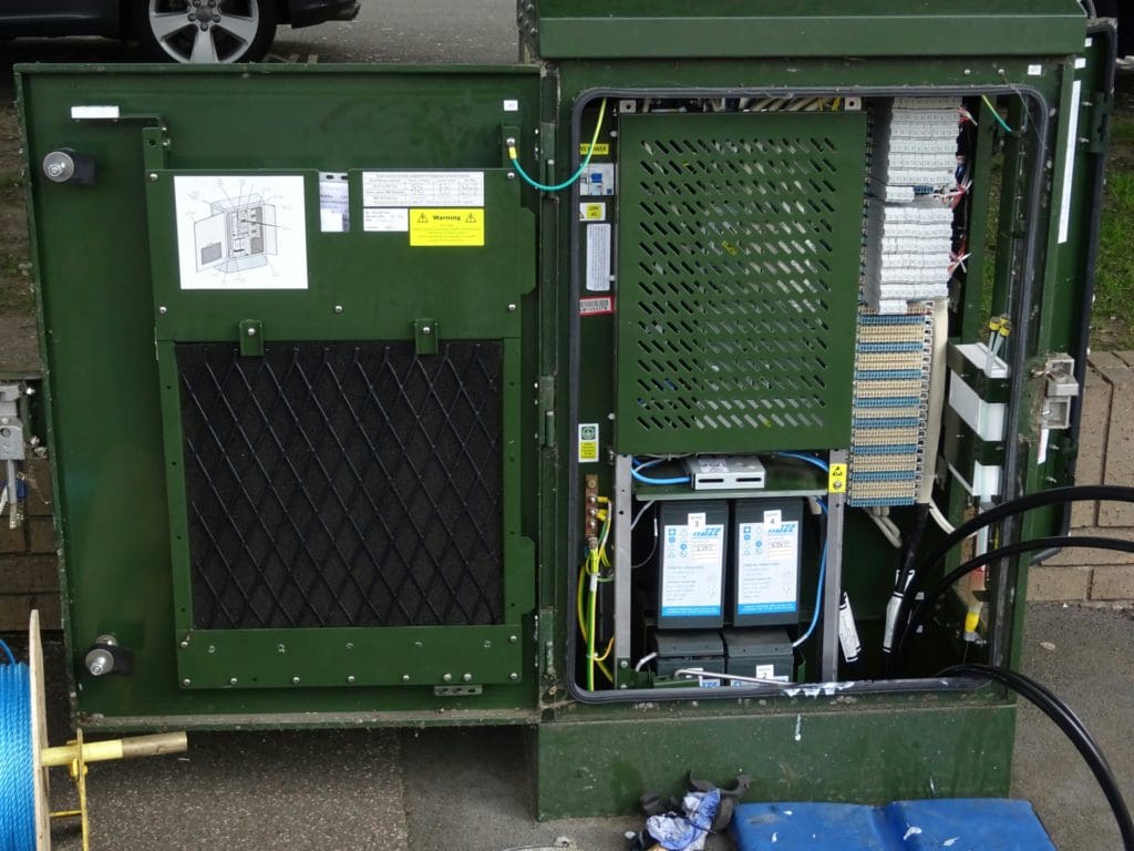 An open green metal cabinet contains various telecommunications equipment, including wiring, cables, and circuit boards. A diagram is attached to the inside of the cabinet door. Tools and wires are scattered on the ground nearby.
