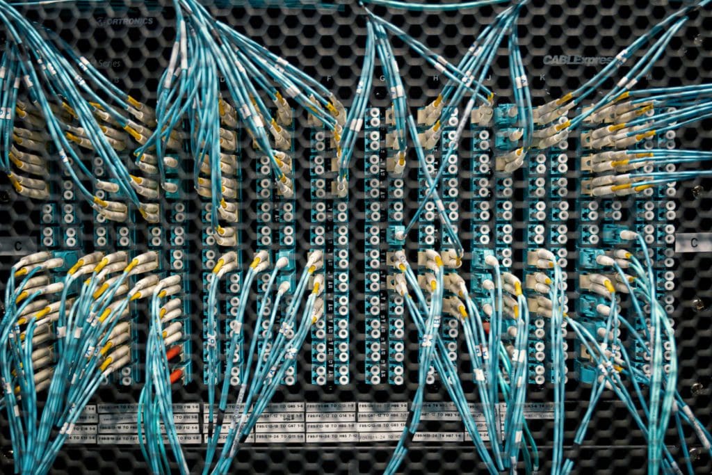 A close-up view of a network server panel with numerous blue and white Ethernet cables connected to multiple ports. The cables crisscross each other, creating a complex, intricate pattern against a dark, perforated background.