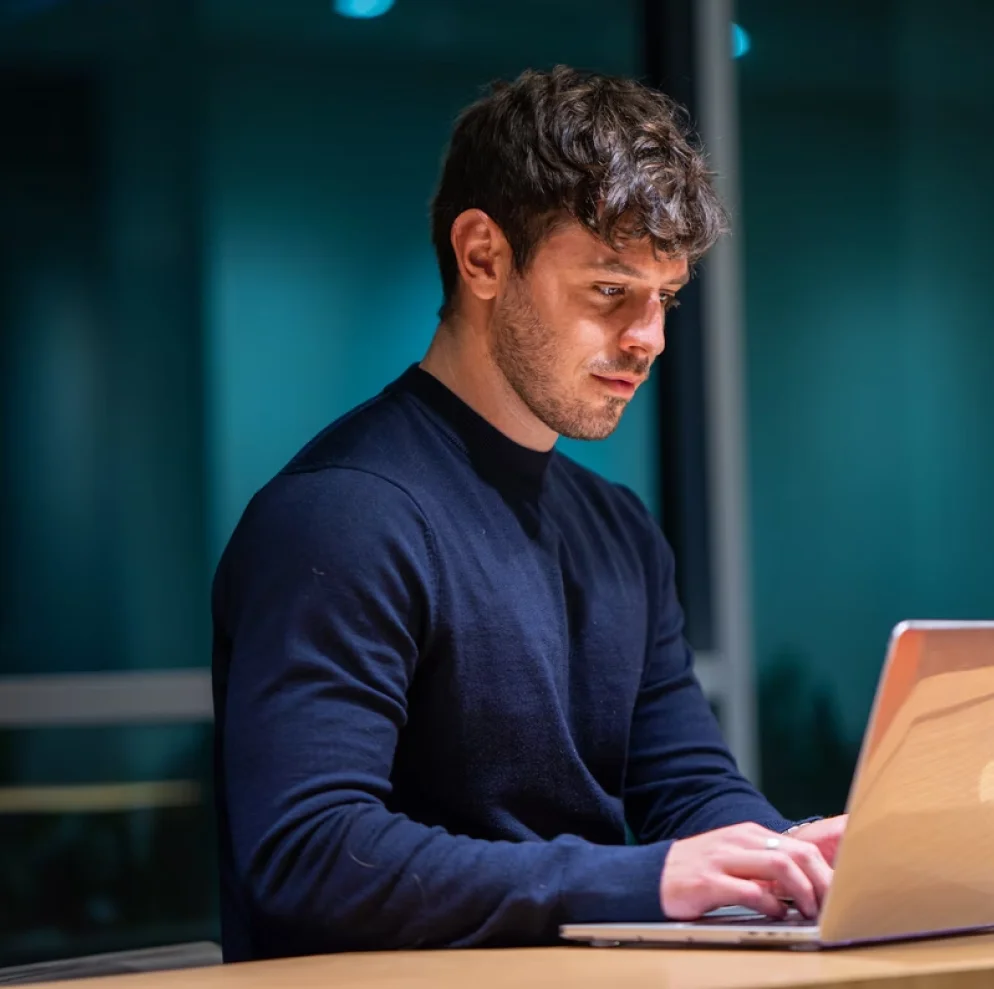 A man with short, dark hair is sitting at a wooden table, typing on a laptop. He is wearing a navy blue long-sleeve shirt and appears focused. The background is slightly blurred with what appears to be a window and some indoor lighting.