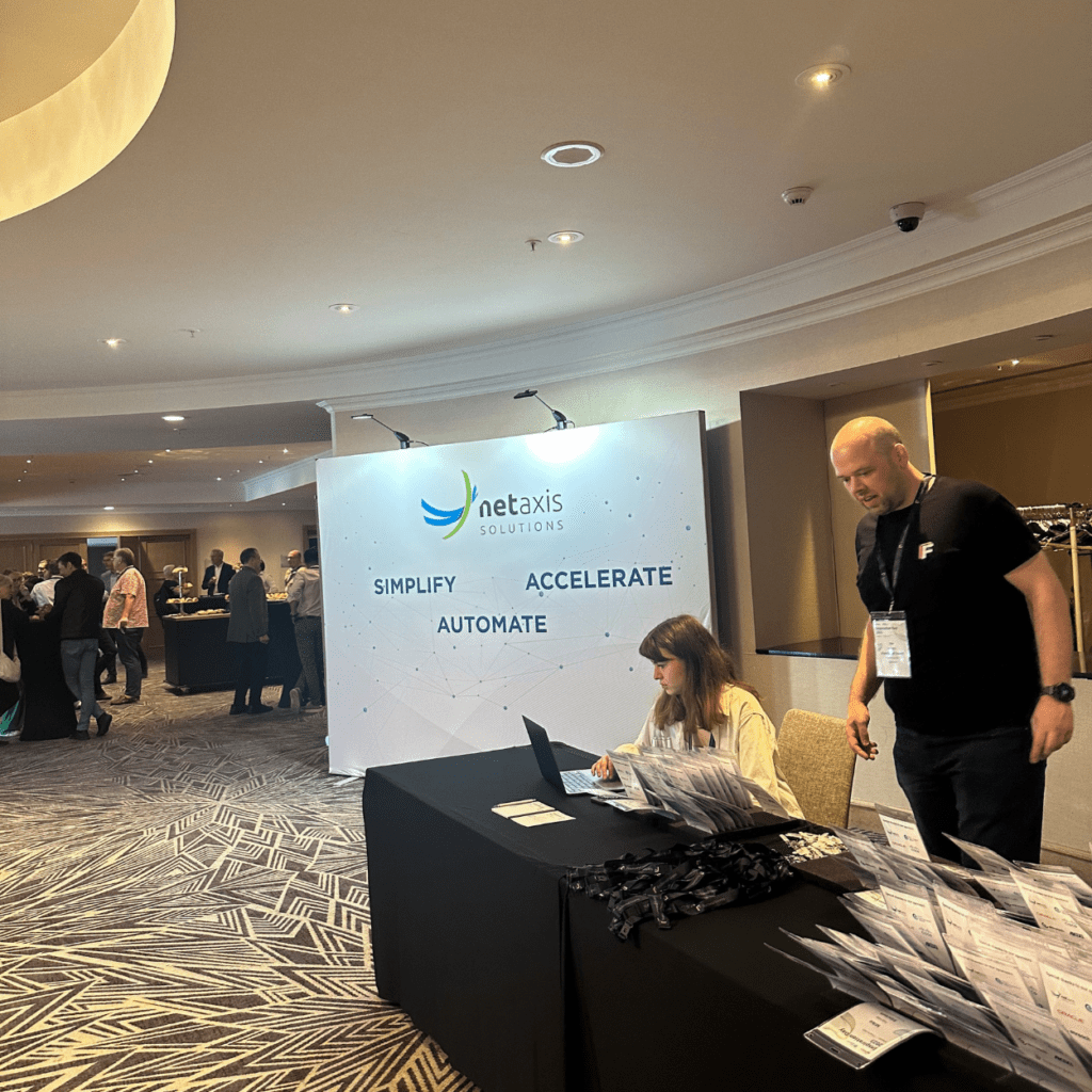 A woman sits at a registration desk with a laptop, surrounded by event materials. A man stands next to the table. Behind them is a large banner with the logo and text of "netaxis solutions" along with "SIMPLIFY," "ACCELERATE," and "AUTOMATE." People are mingling in the background.