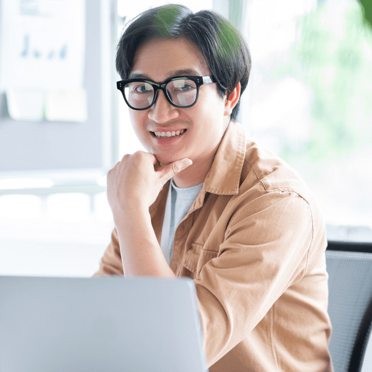 A man dressed in a button-up shirt sits at a desk in an office. He is smiling in front of his laptop.